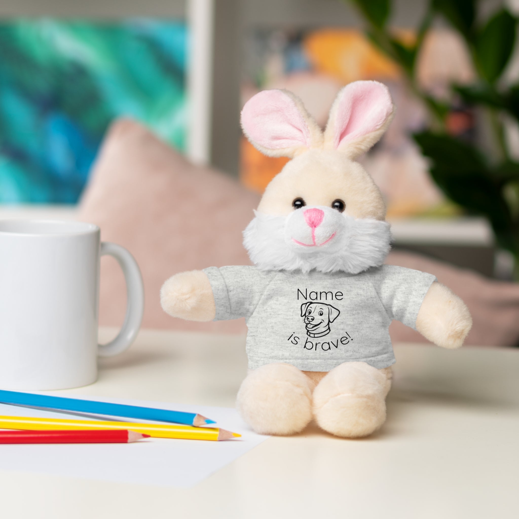Soft off white plush bunny wearing a Name is Brave personalized grey  tee shirt - sitting on desk with colored pencils
